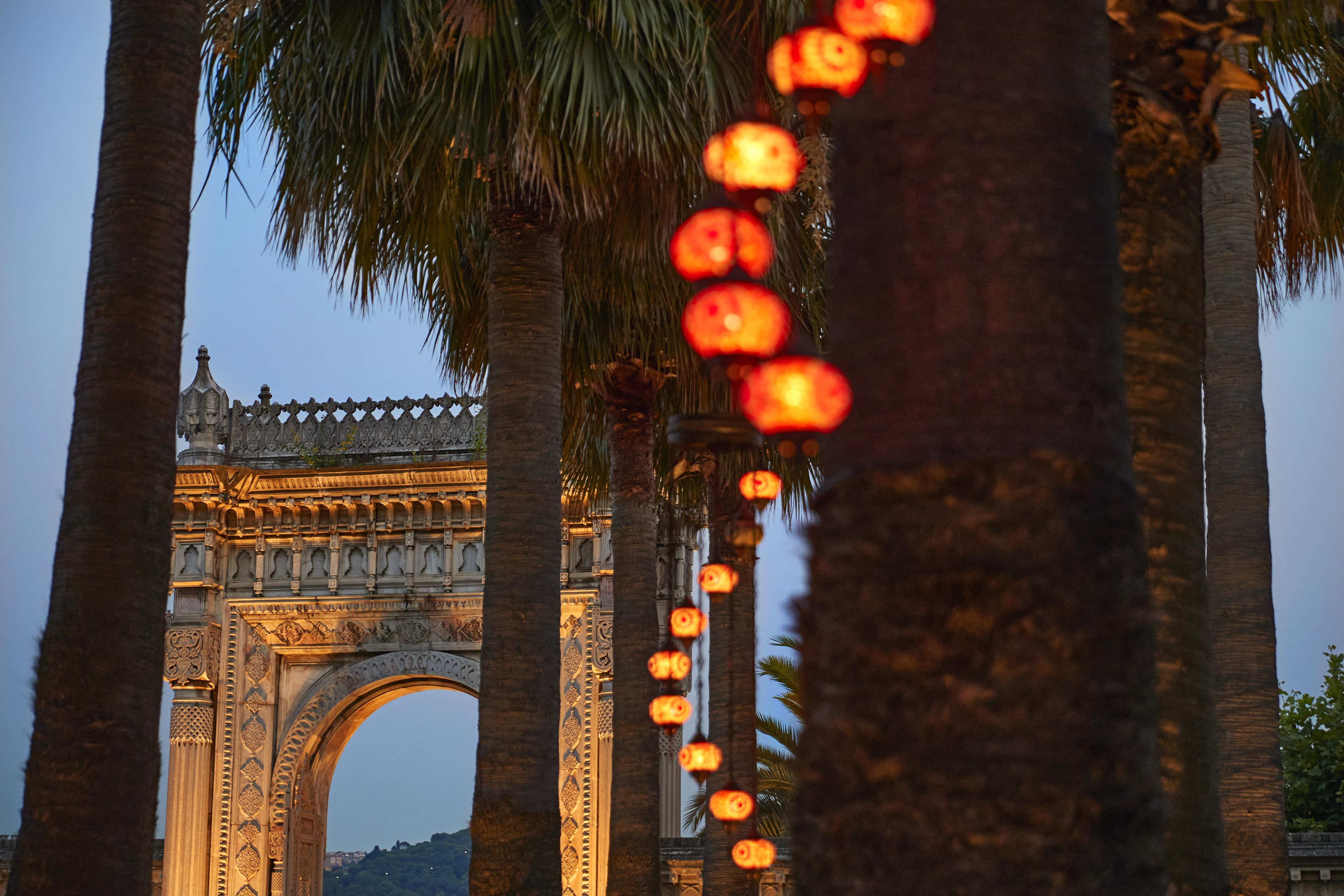 Un arco histórico con intrincados grabados, rodeado de palmeras e iluminado con luces colgantes.