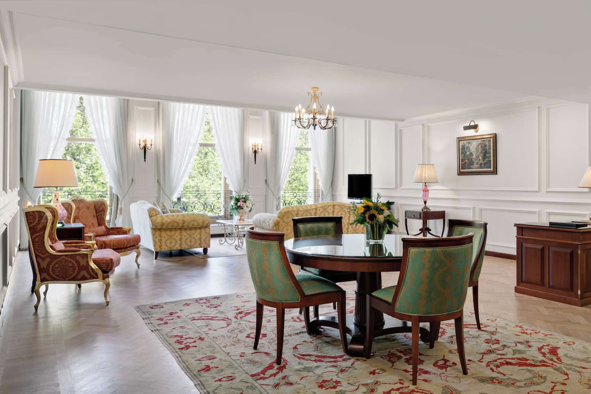 A spacious living room with a dining area, featuring a chandelier, comfortable seating, and a large window with curtains.