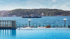 A waterfront view with a ferry, a pool with lounge chairs, and a hill with buildings in the background. - Ciragan Palace Kempinski (Istanbul)