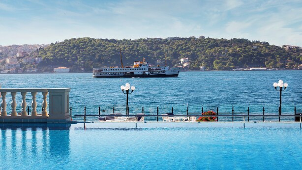 Vista al agua con un ferry, una piscina con sillones de descanso y una colina con edificios al fondo.