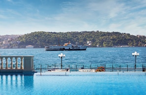 A waterfront view with a ferry, a pool with lounge chairs, and a hill with buildings in the background. - Ciragan Palace Kempinski (Istanbul)