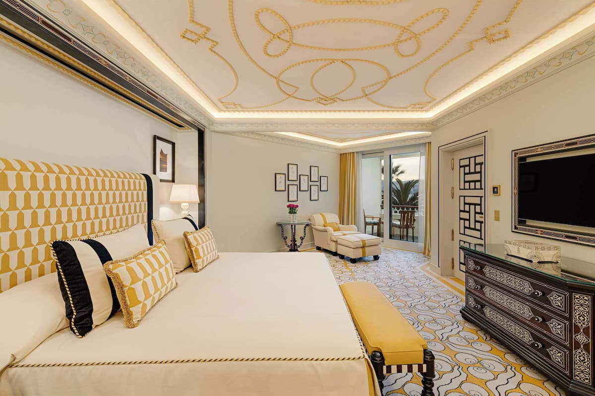 A hotel room with a large bed, a TV, a dresser, and a seating area with a chair and ottoman.
