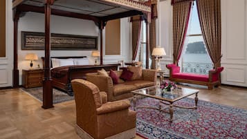 Un dormitorio lujoso con cama con dosel, una sala de estar con sofá y sillones, una mesa de centro de vidrio, y vista a una masa de agua a través de amplios ventanales.