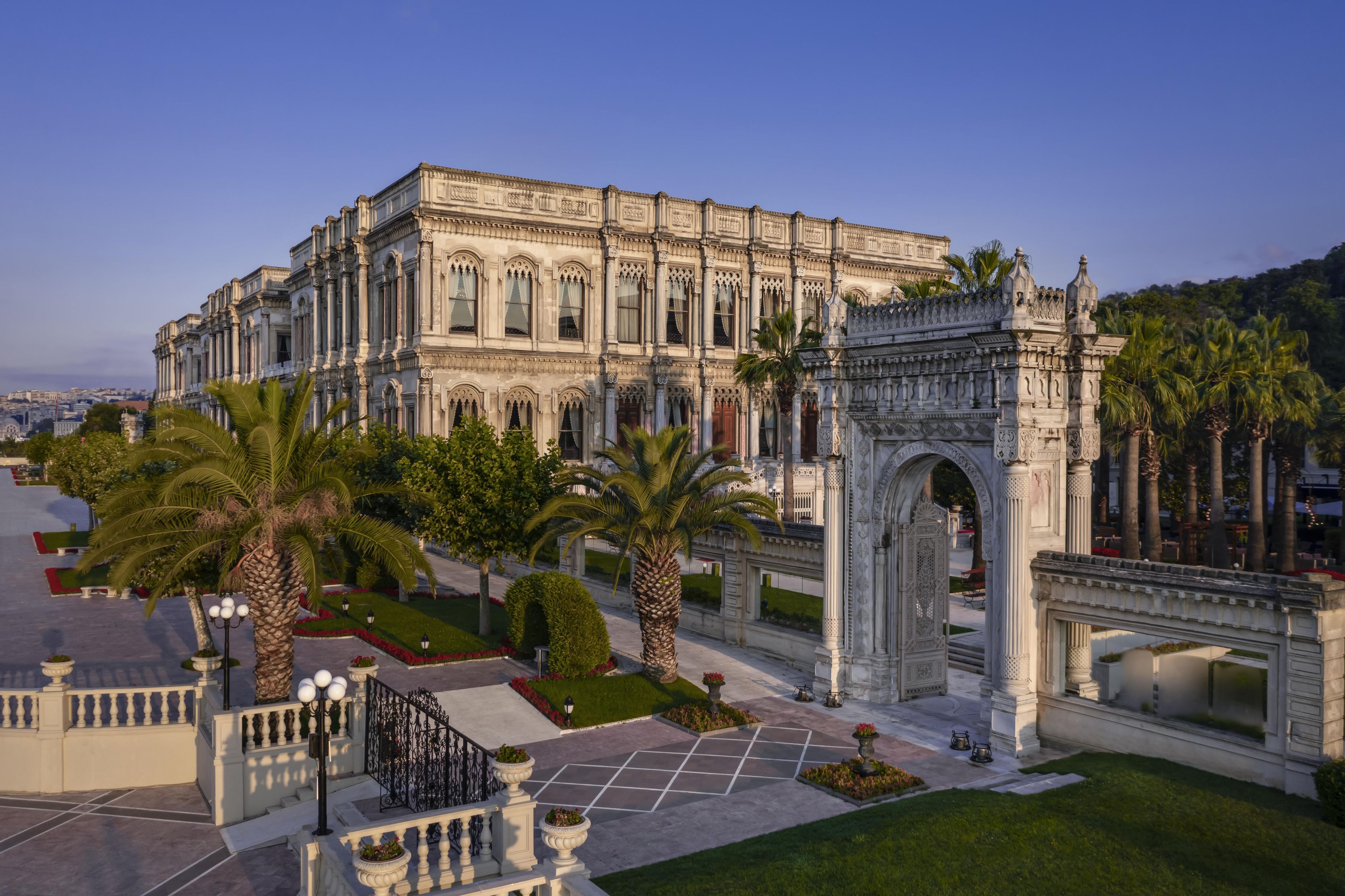 Un grand bâtiment somptueux, doté d’une architecture classique, avec des colonnes et une arche, entouré de jardins bien entretenus et de palmiers.