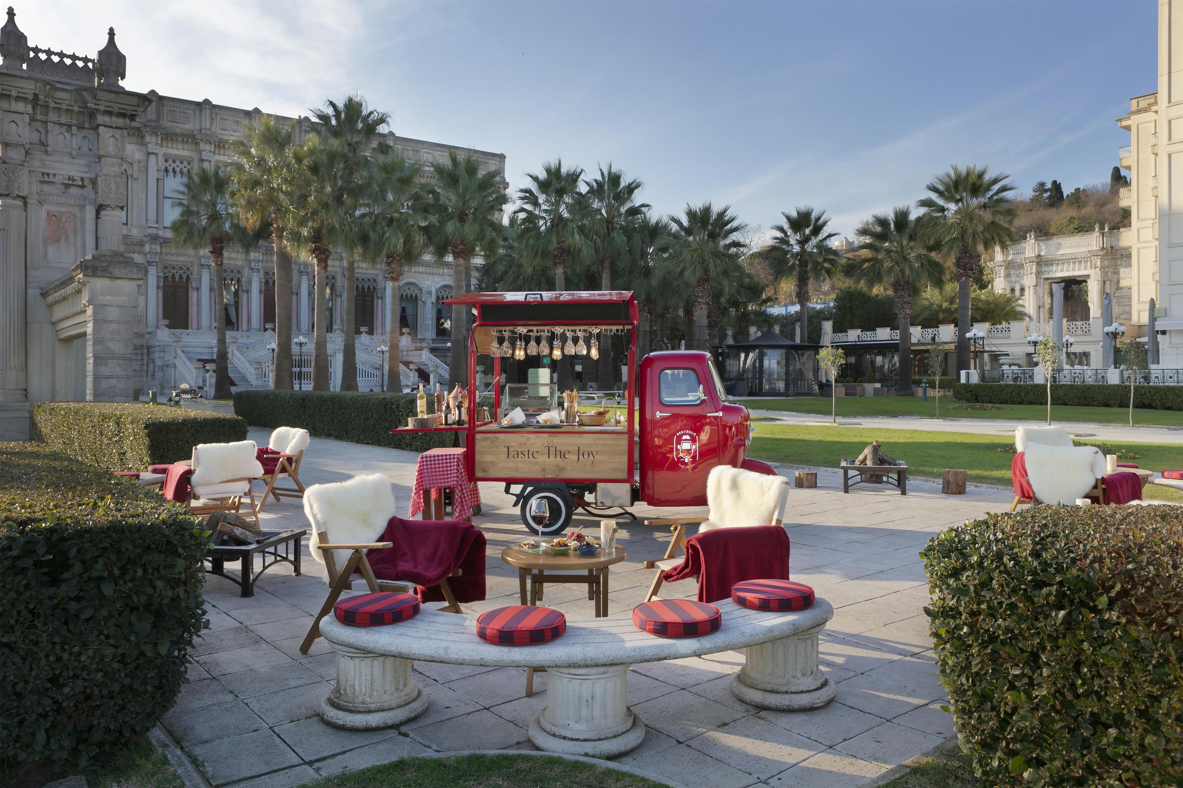 A vintage red food truck with a sign reading 'Taste The Joy' parked in a courtyard with outdoor seating and palm trees.