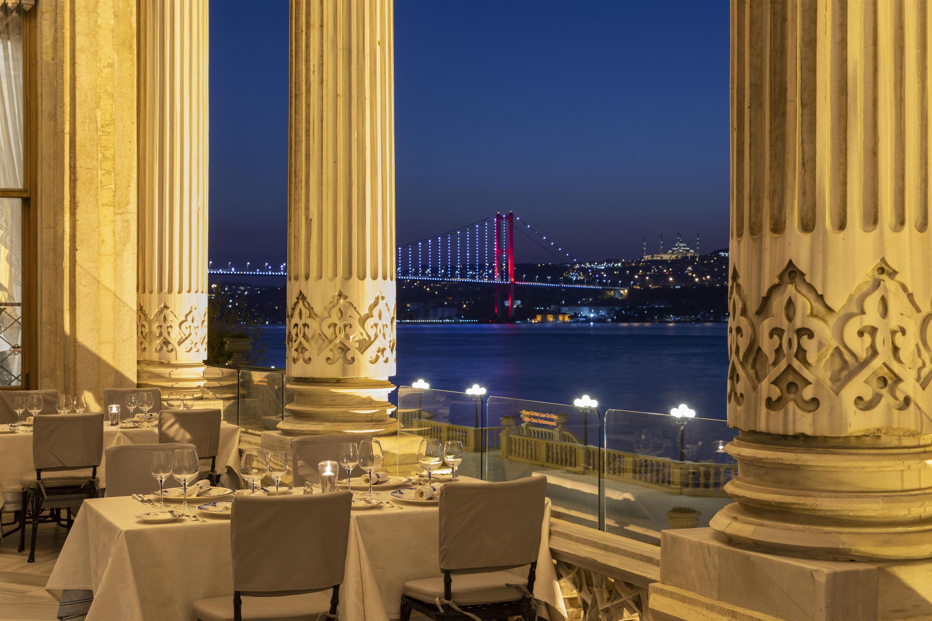 Un restaurant avec des tables dressées pour le service, donnant sur une étendue d’eau, avec un pont en arrière-plan.