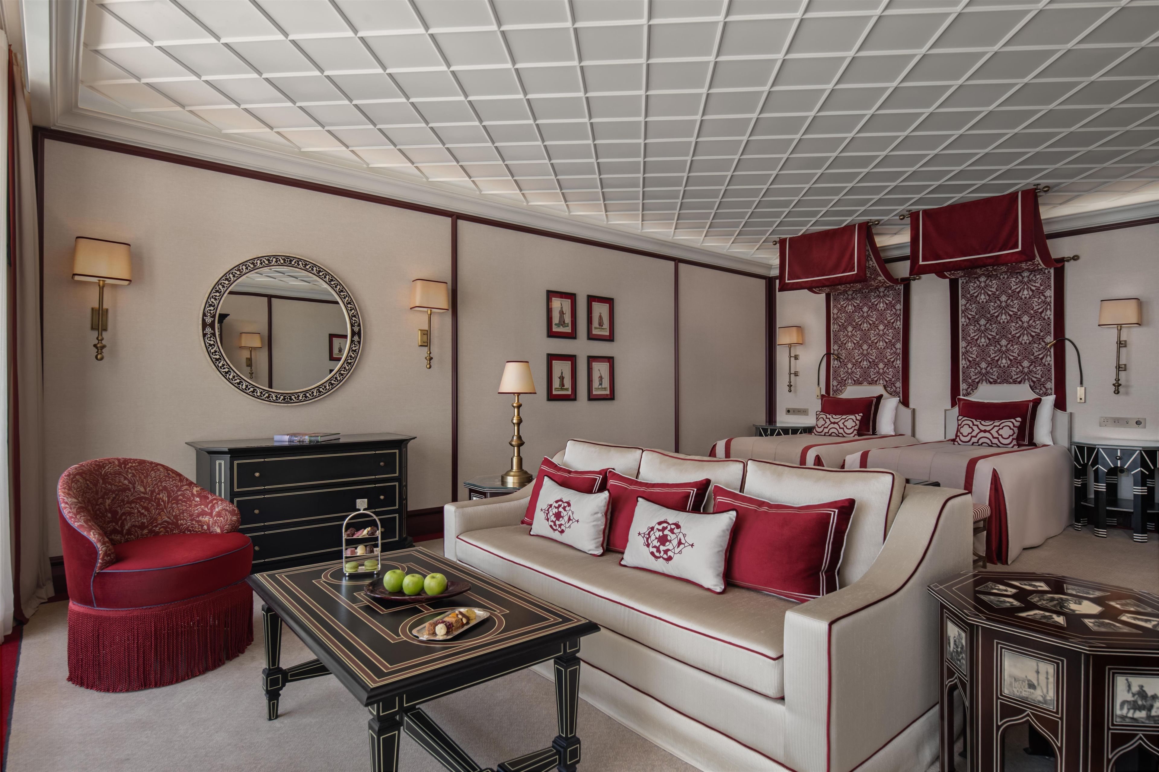 Habitación de hotel con una cama grande, un sofá, una mesa de comedor con un centro de mesa decorativo, un sillón rojo y un espejo en la pared.
