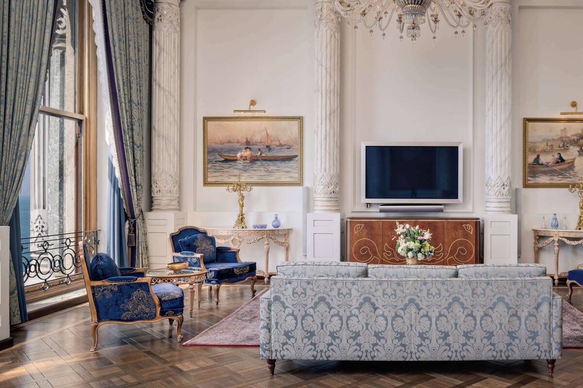A luxurious living room with a large sofa, ornate chairs, a television, and classical paintings.