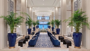 Un vestíbulo de hotel con un mostrador de recepción alargado, asientos tapizados en azul, grandes plantas en macetas y una alfombra estampada.