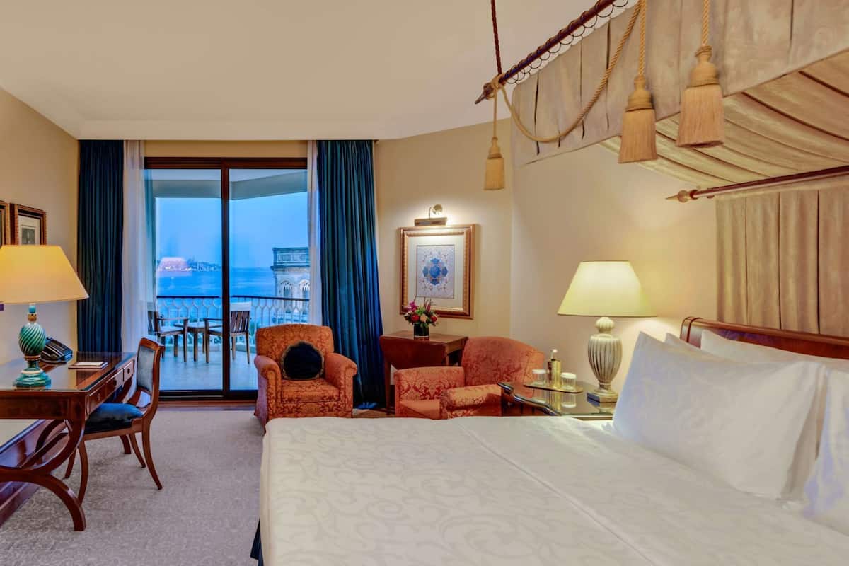 A hotel room with a bed, two armchairs, a desk, a chair, a nightstand, a lamp, a picture frame, and a view of the sea through a large window.