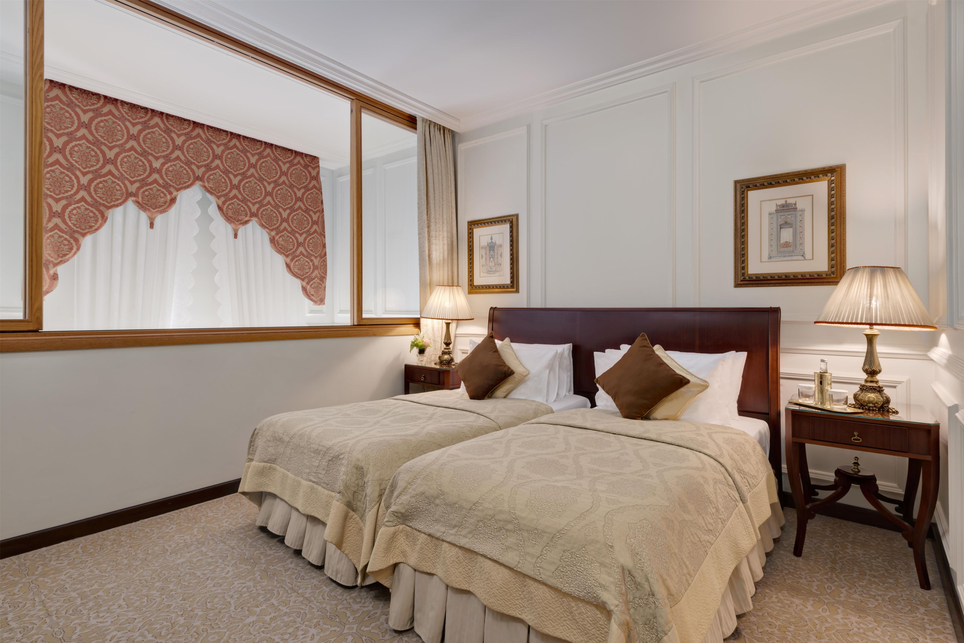 Habitación de hotel con dos camas, un ventanal con cortinas, una mesita de noche con lámpara y un cuadro en la pared.