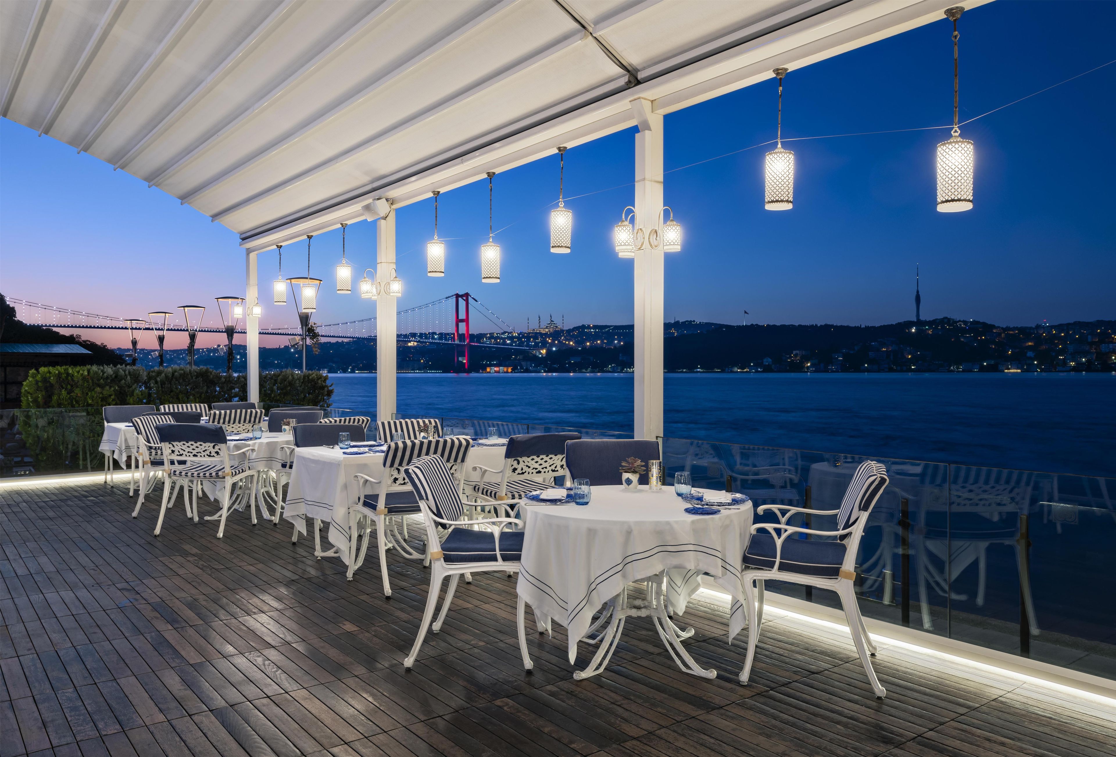 An outdoor dining area with tables and chairs, overlooking a body of water with a bridge in the background.