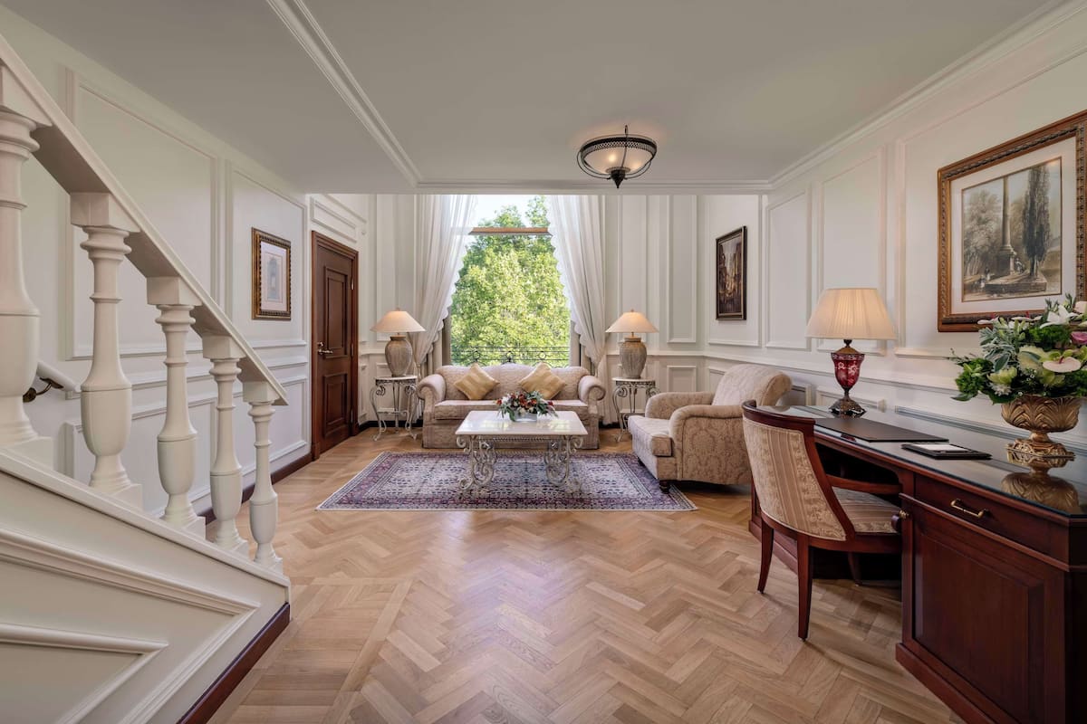 A spacious living room with a herringbone floor, a large window with a view of trees, a staircase with white balusters, and a desk with a lamp and vase.