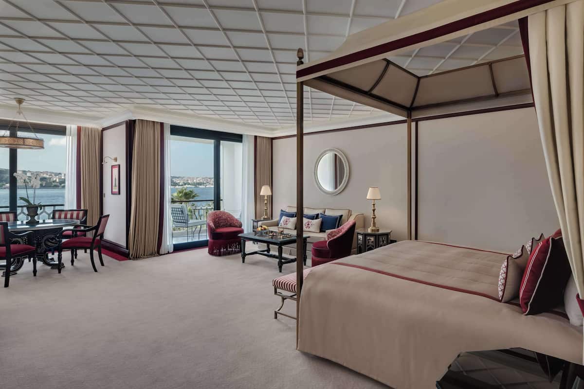 A hotel room with a four-poster bed, a dining area with a table and chairs, a sofa, and a view of the sea.