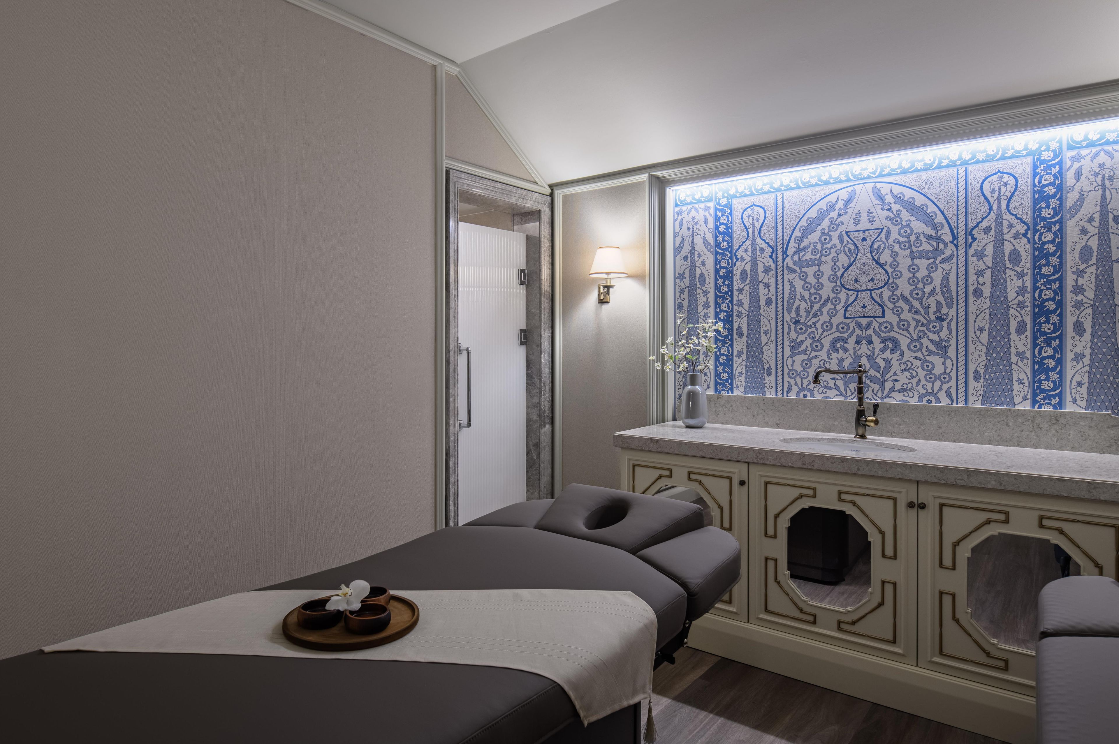 Una habitación con cama, mesa de masajes, lavamanos y un detalle decorativo en la pared.