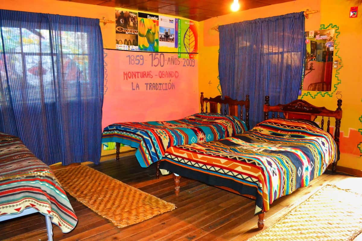 Une chambre avec deux lits en bois, des couvertures colorées, un tapis tissé et un mur orné d’une frise chronologique retraçant des événements historiques.