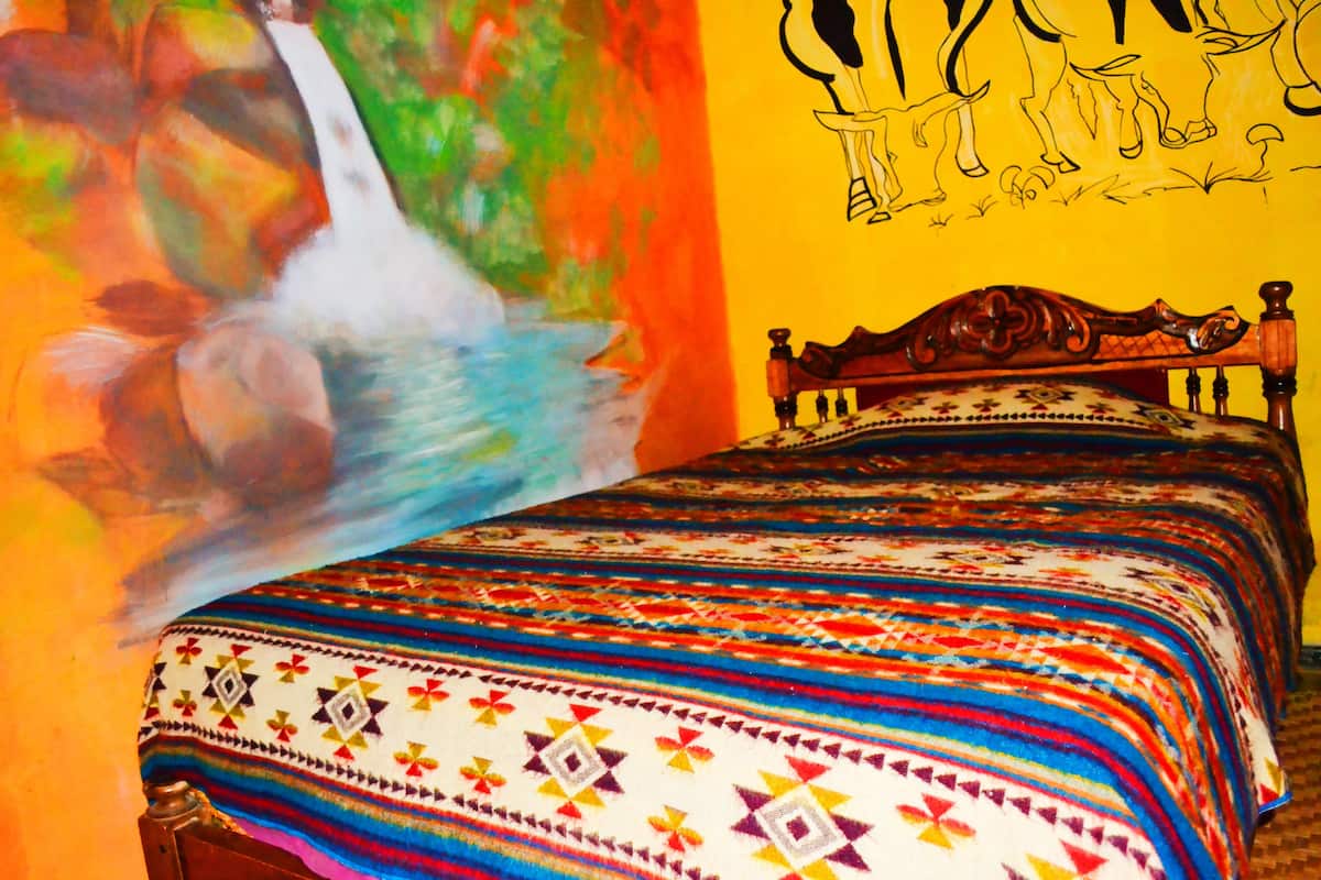 Un lit recouvert d’une couverture colorée et un mur orné d’un tableau représentant une cascade et des chevaux.