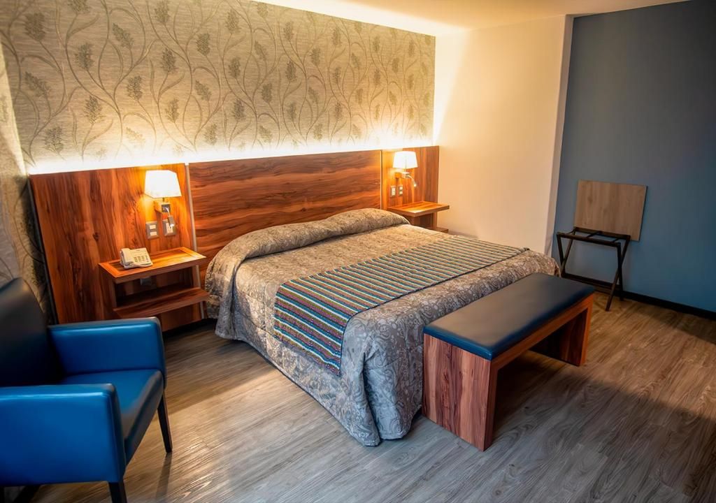 A bedroom with a large bed, two bedside lamps, a wooden headboard, a bench, and a chair.