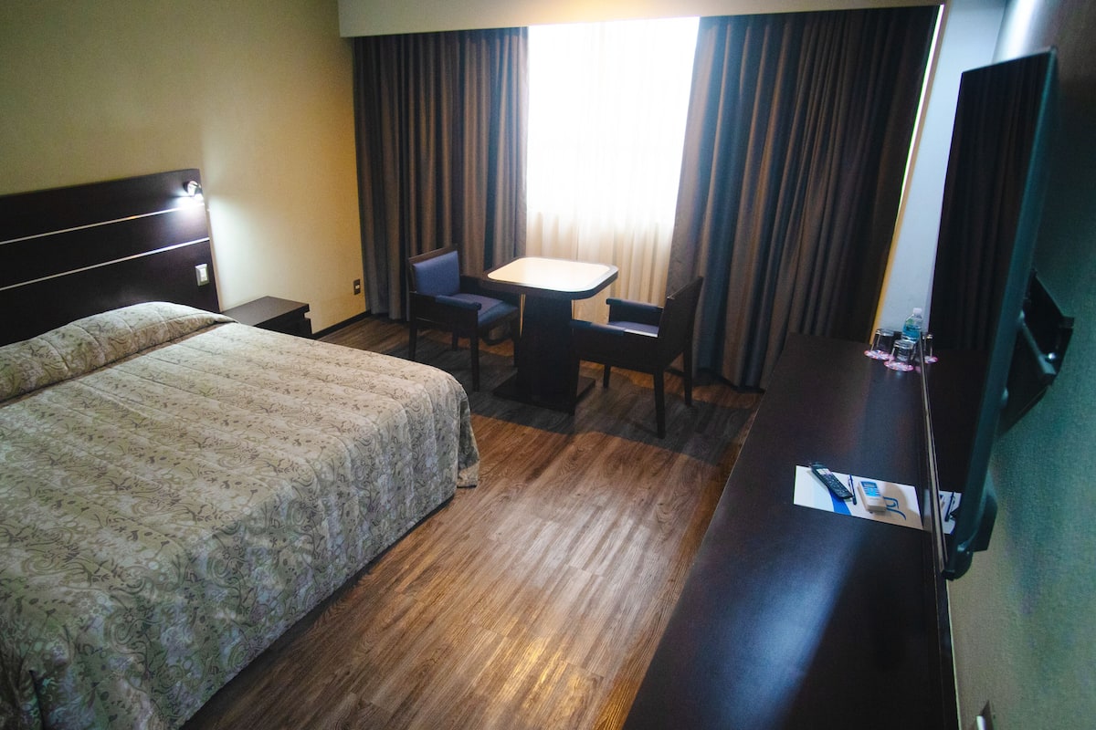 A hotel room with a large bed, a small table, two chairs, a TV, and a window with curtains.