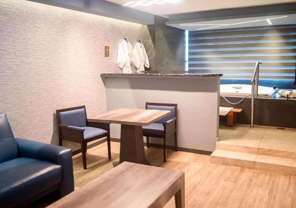 Habitación de hotel moderna con piso de madera, zona de estar con mesa y sillas, kitchenette pequeña y baño con cortina de ducha a rayas azules.