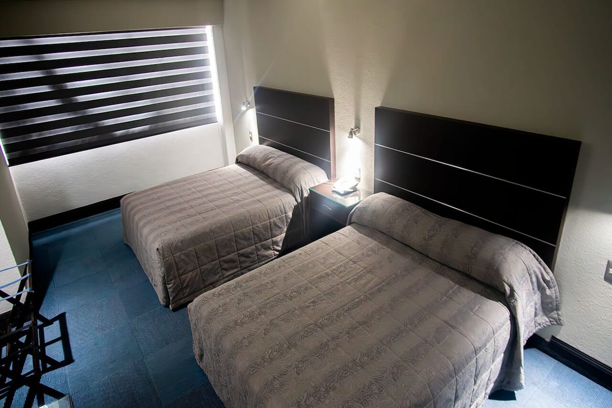 A hotel room with two beds, a window with blinds, a bedside table with a lamp, and a blue carpet.