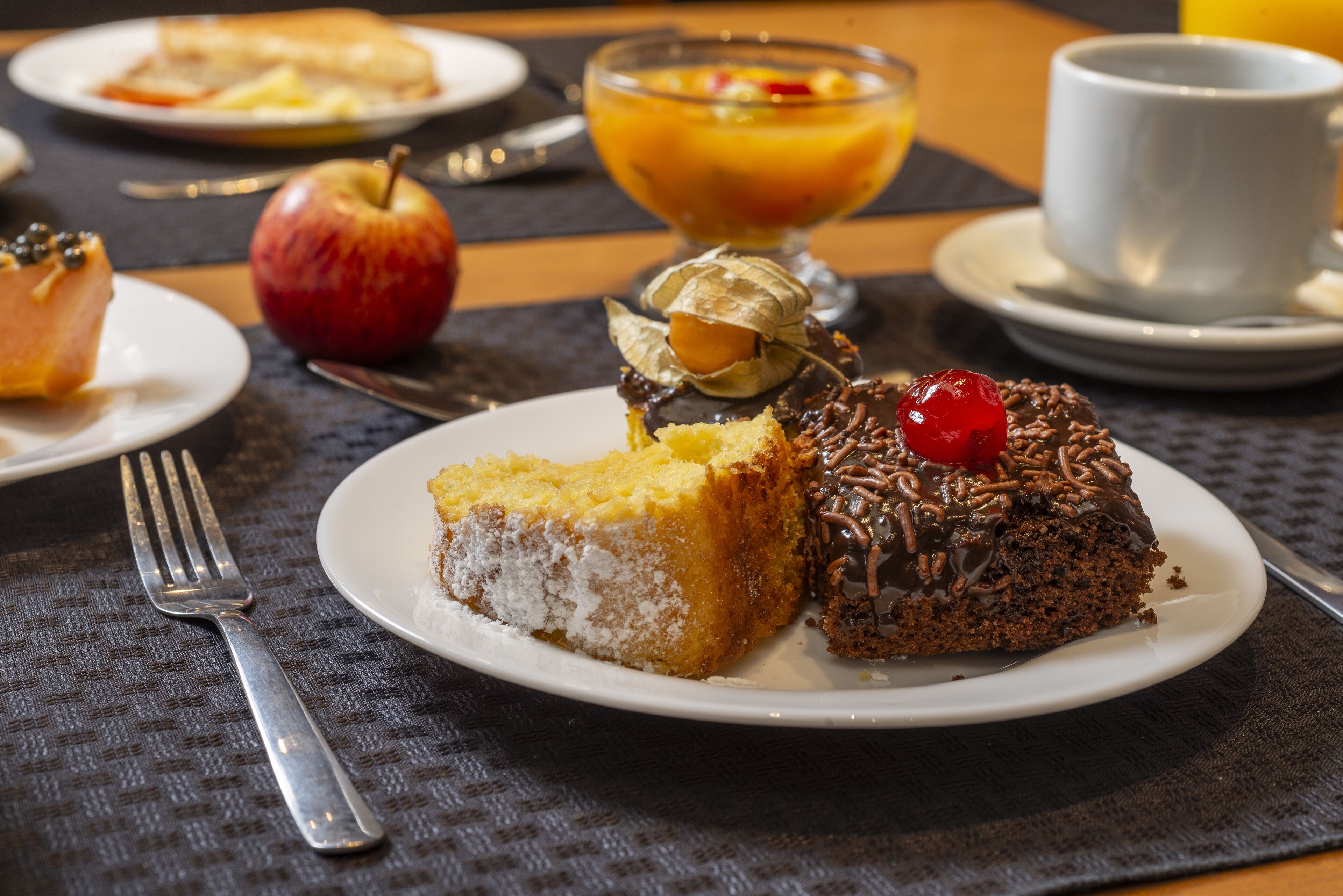 A dining table with plates of cake, a bowl of fruit, and a cup of coffee.