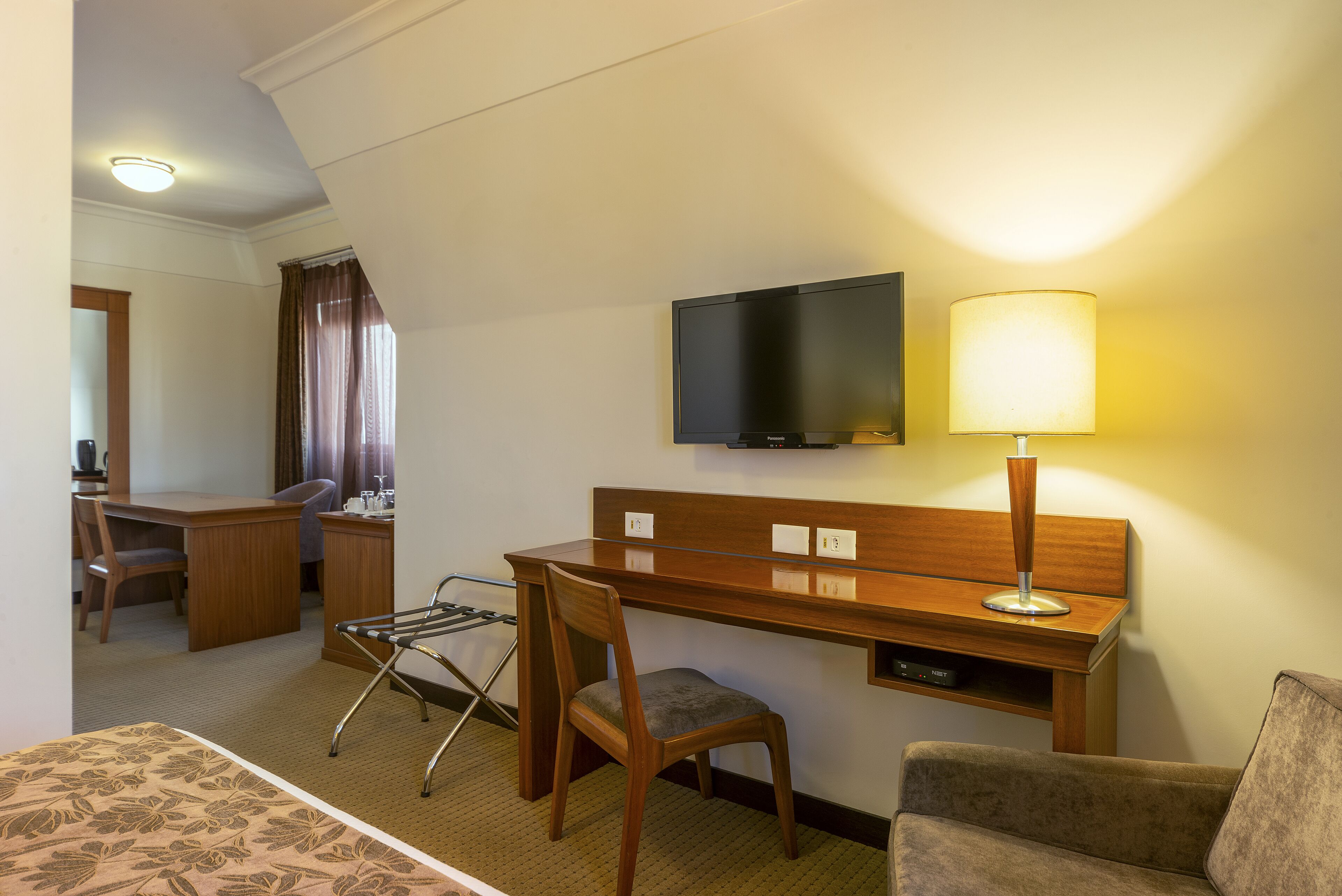 A hotel room with a wooden desk, a television, a lamp, a chair, and a bed.