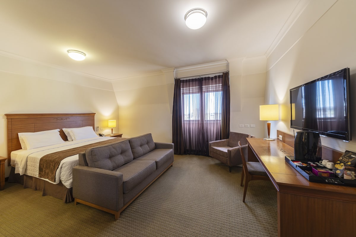 Une chambre d’hôtel équipée d’un lit, d’un canapé, d’un bureau, d’une chaise, d’une télévision et d’une fenêtre avec des rideaux.