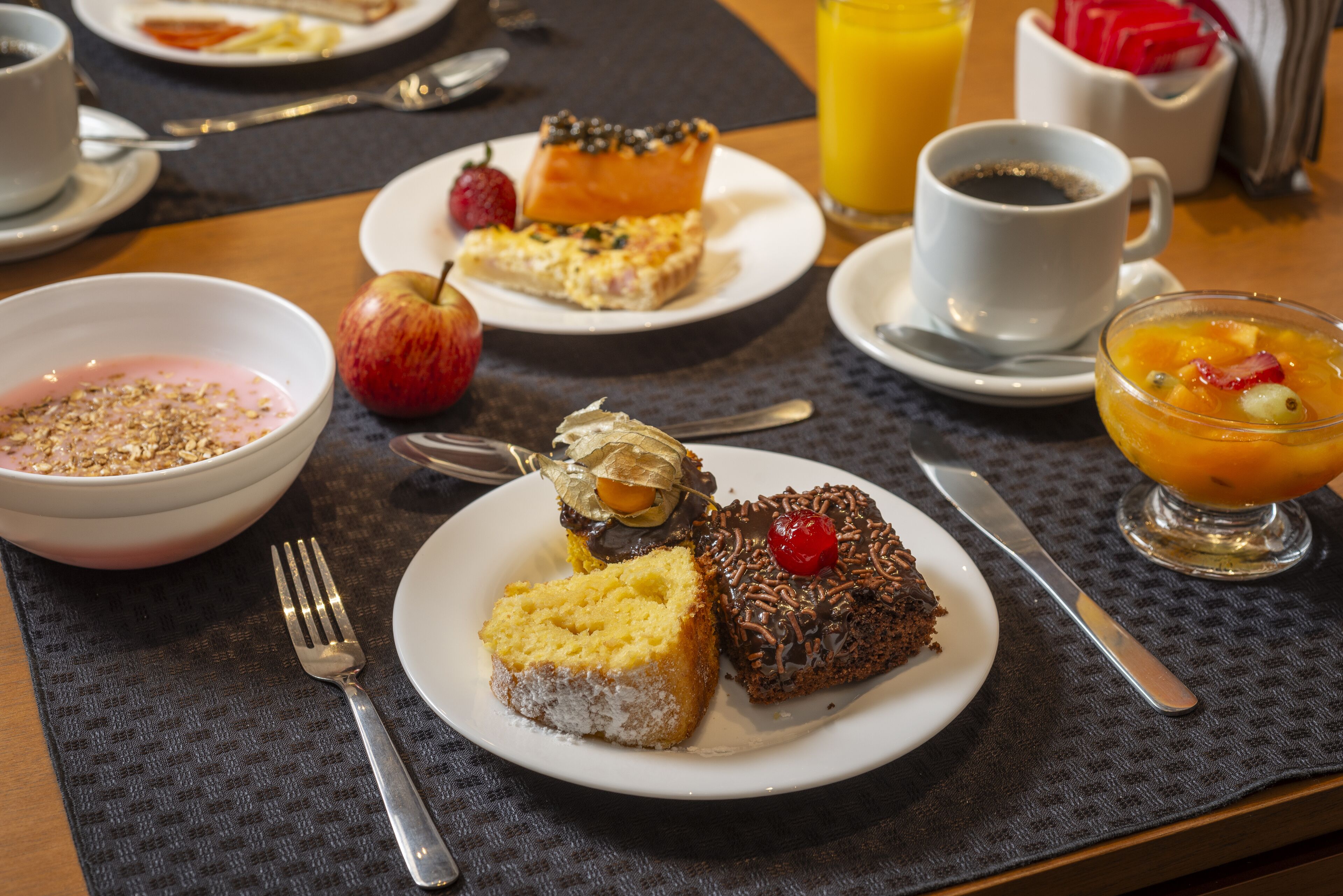 A breakfast setting with various dishes including a bowl of cereal, a slice of cake, and a glass of orange juice.