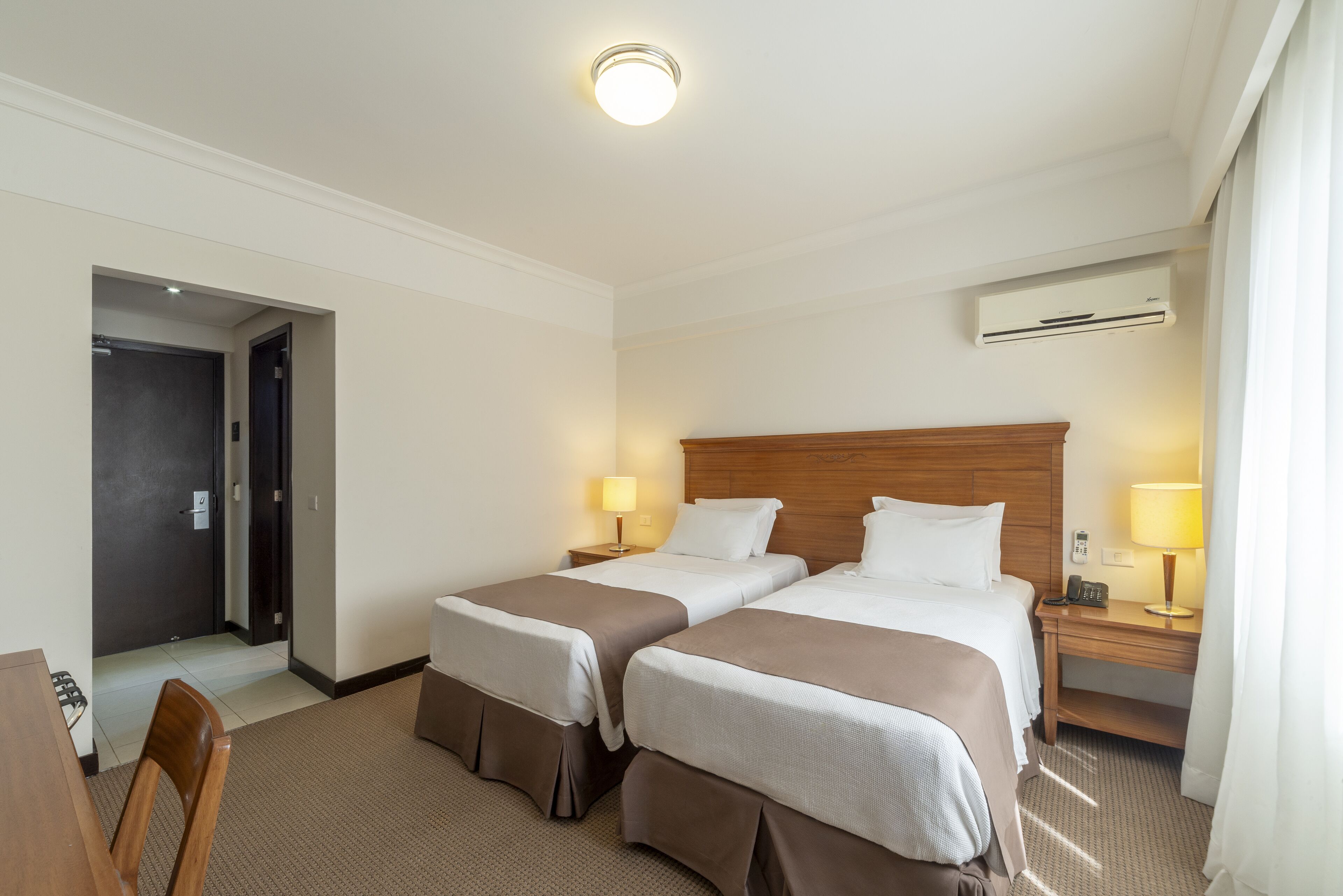 A hotel room with two beds, a desk, a chair, a door, and an air conditioning unit.