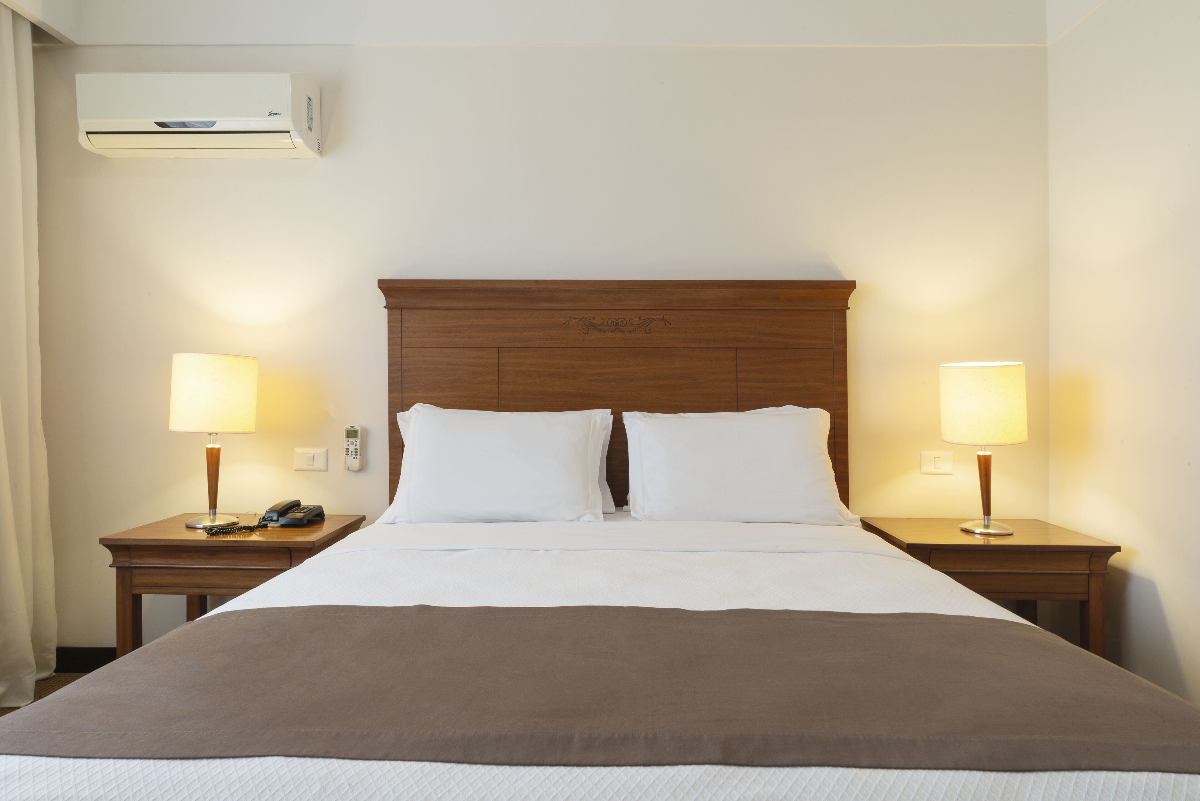 A hotel room with a large bed, two bedside tables with lamps, a telephone, and an air conditioning unit.