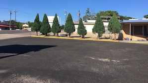 Un aparcament amb una filera d'arbres de fulles perennes, un edifici amb un rètol i un cel blau clar.