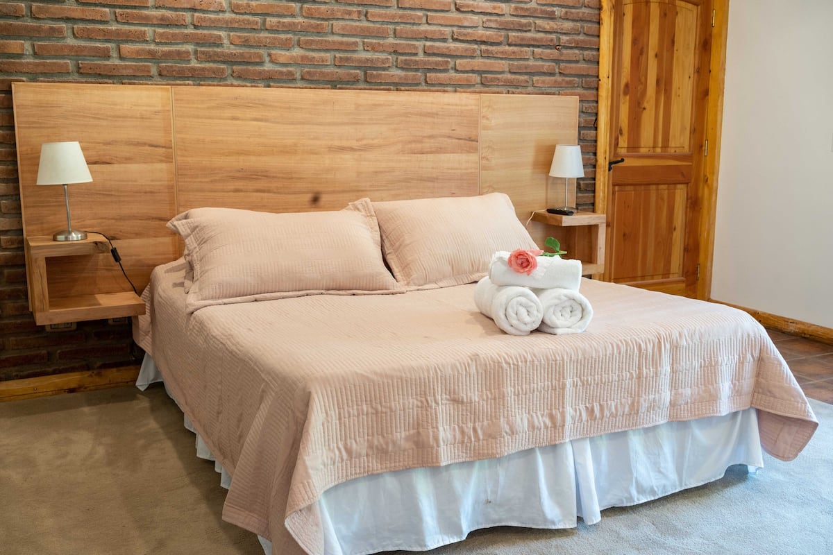 Una cama bien tendida con un edredón de color claro y almohadas, un cabecero de madera y una mesita de noche con una lámpara.