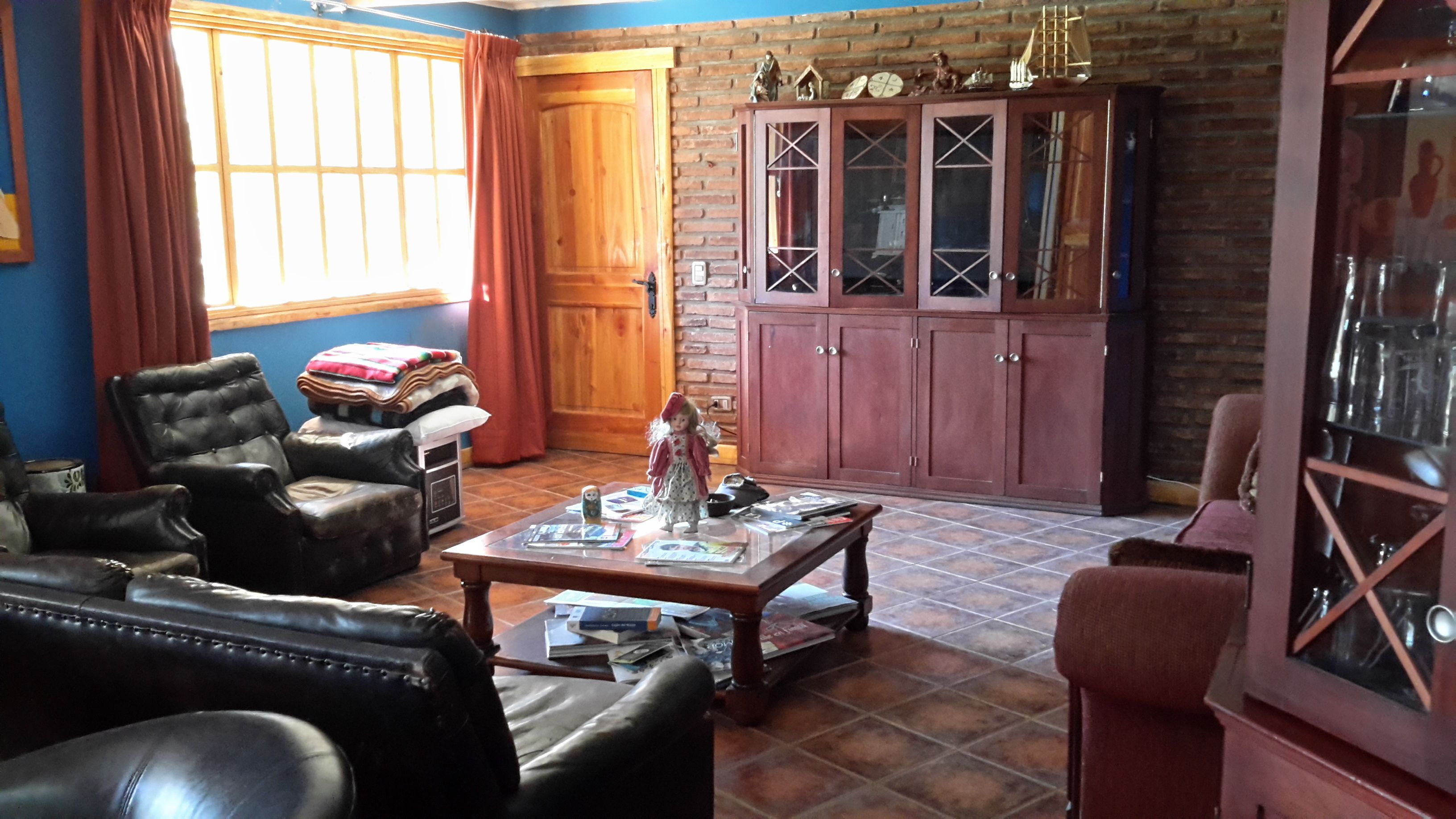 Una sala de estar con pared de ladrillo, un aparador de madera con puertas de cristal, un sillón de cuero, una mesa de centro con libros y una ventana con cortinas.