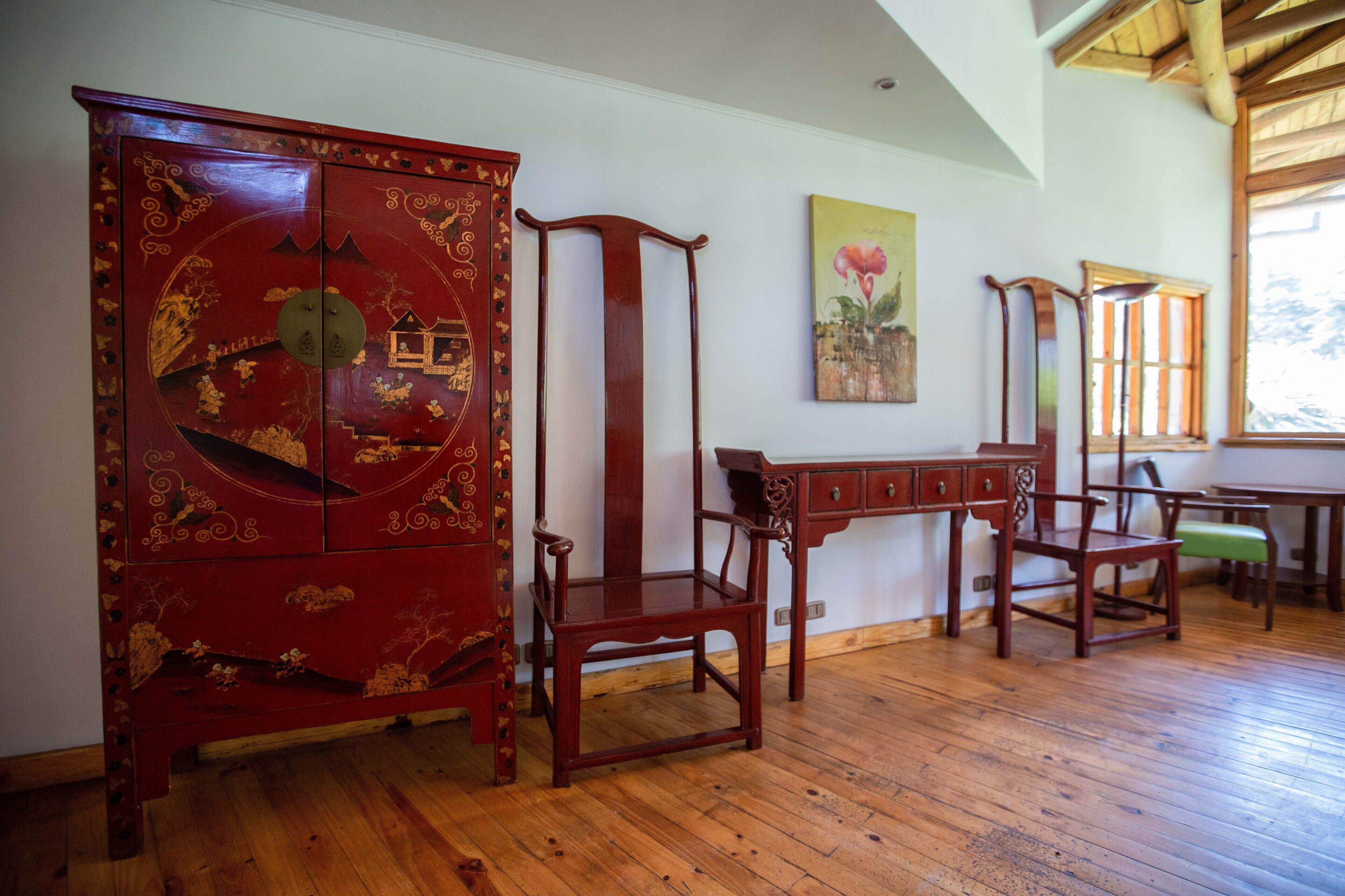 Una habitación con piso de madera, un aparador tradicional rojo con diseños intrincados, una silla de madera, una mesita y un cuadro en la pared.