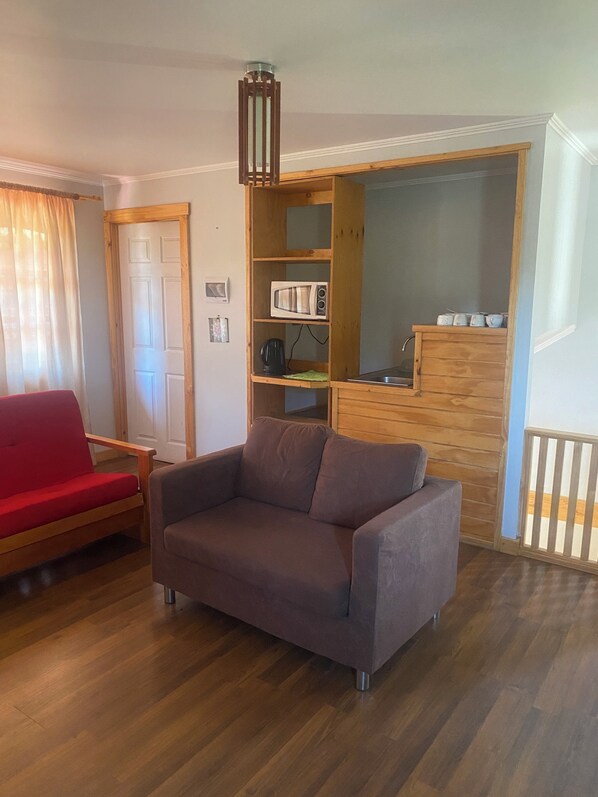 A living room with a brown sofa, a red chair, a wooden cabinet with kitchen appliances, and a wooden floor. - La Casa de Carlitos (San José de Maipo)