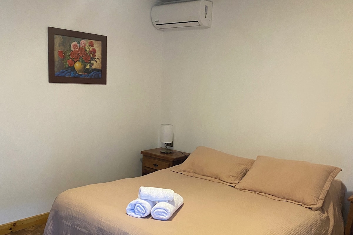 Una cama bien hecha con una colcha color beige y almohadas, una mesita de noche con una lámpara y un equipo de aire acondicionado en la pared.