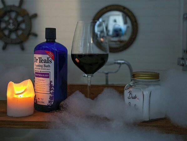 A bottle of Dr. Teal's Foaming Bath, a lit candle, a glass of red wine, and a jar labeled "Bath" on a wooden surface.