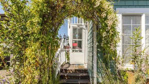 A wooden staircase leading to a white door with a window, surrounded by lush greenery and flowering plants. - Enchanted Cottages (Seaview)