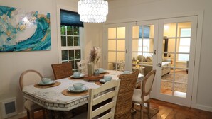 A dining area with a round table, chairs, and a chandelier. There is a window with blinds and a door leading to another room. - Enchanted Cottages (Seaview)