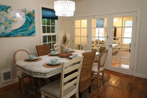A dining area with a round table, chairs, and a chandelier. There is a window with blinds and a door leading to another room.
