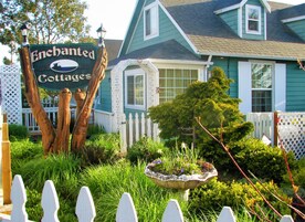 Un letrero para Enchanted Cottages, con cerca de piquetes blancos, un abrevadero para pájaros y abundante follaje.