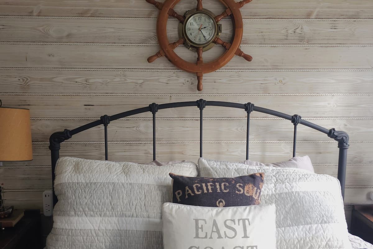 Ein Bett mit einer gestreiften Tagesdecke, zwei Kissen, eines mit der Aufschrift "PACIFIC SOUTH EAST COAST" und einer Schiffsschleuderuhr an der Wand.
