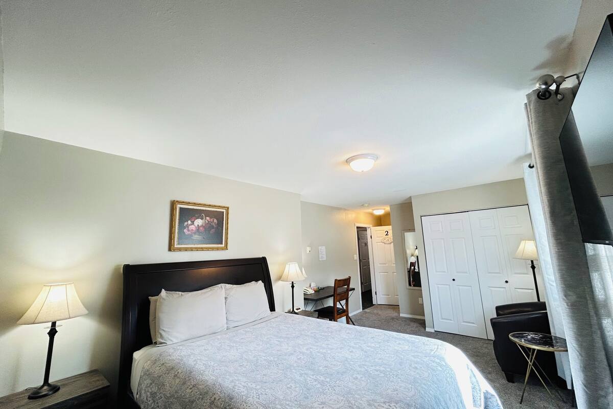A hotel room with a bed, bedside lamps, a framed picture on the wall, a door, and a small table with a chair.