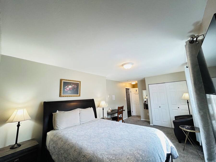 Quarto de hotel com cama, luminárias de cabeceira, um quadro na parede, uma porta e uma pequena mesa com uma cadeira.