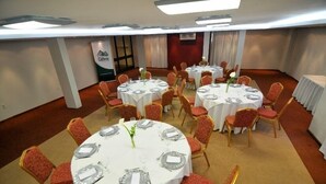 Un salón de conferencias con varias mesas redondas, cada una con mantel blanco, sillas tapizadas en rojo y cubiertos.