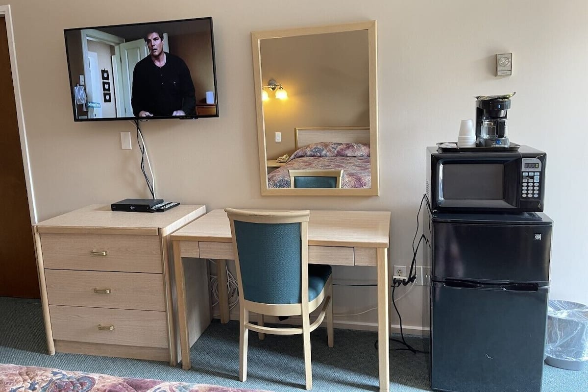 A hotel room with a desk, chair, television, mirror, and a refrigerator with a microwave on top.