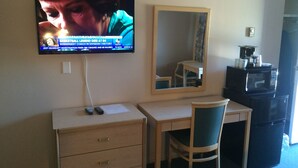 A hotel room with a TV mounted on the wall displaying news, a wooden desk with a chair, a mirror, and a microwave on a cabinet. - Rider's Inn (Kamloops)