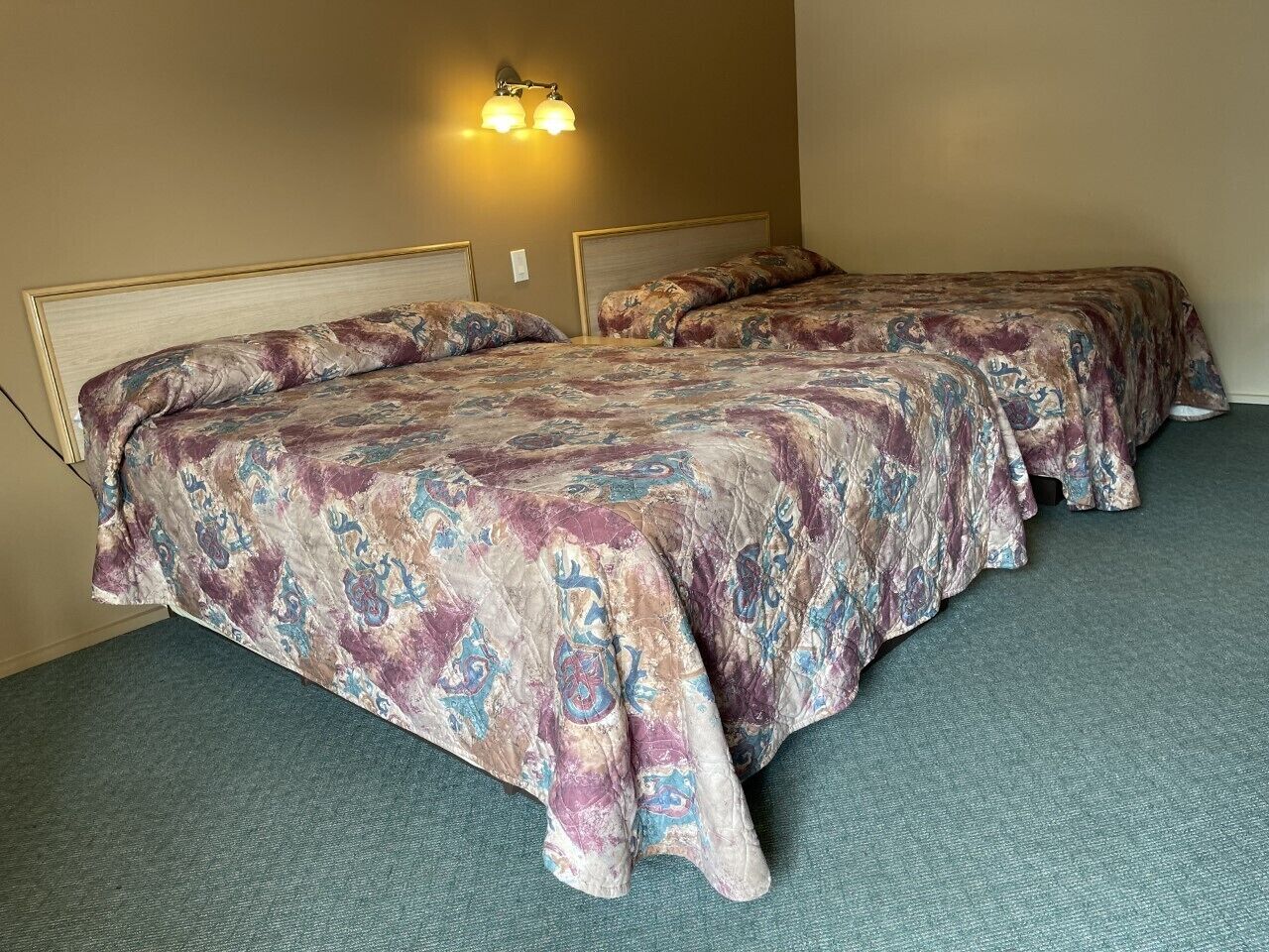 Quarto de hotel com duas camas, piso coberto por carpete e luminária fixada na parede.