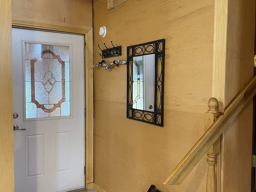 A wooden interior with a white door, a small window, a wall-mounted mirror, and a wooden staircase.